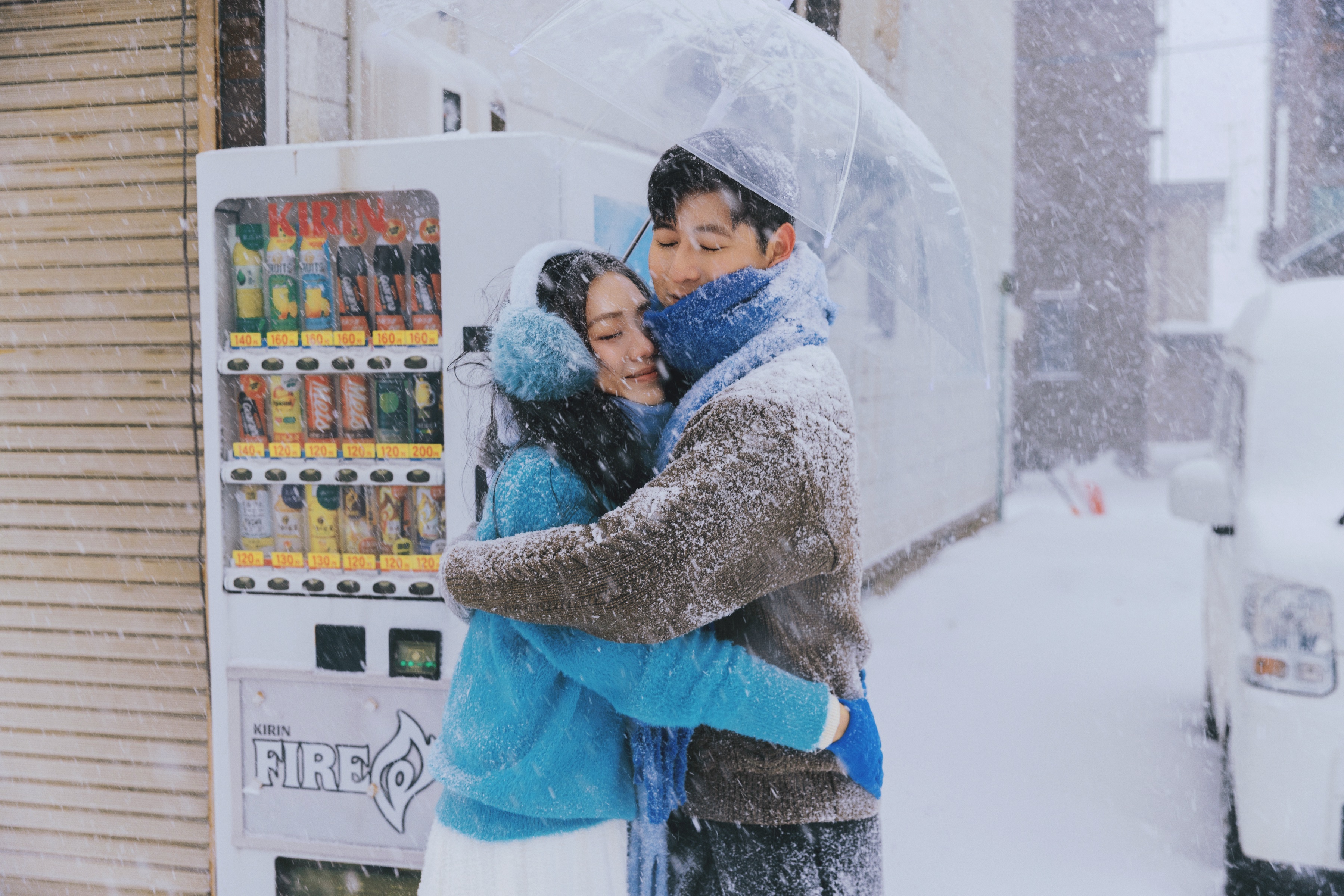 自动售货机旁，撑着透明伞的两个人在雪中相拥